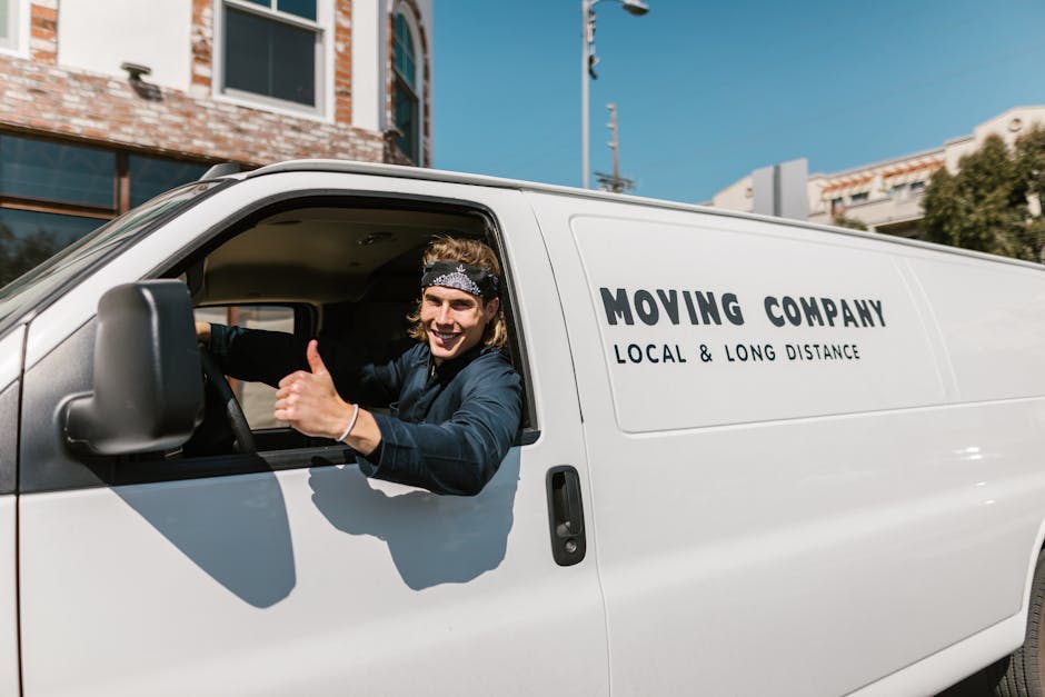 A young male mover with styled hair, wearing a bandana and dark clothing, is seated inside a white moving van, visible through the open driver's side window. He is smiling and giving a thumbs-up gesture. The side of the van displays the text 'MOVING COMPANY' and 'LOCAL & LONG DISTANCE' in black lettering, indicating a professional relocation service. The background shows a clear blue sky, urban buildings with brick and glass facades, and some trees, suggesting an outdoor setting in an urban area during daytime. The van is parked near a building with a brick lower section and large windows, consistent with typical house removal or moving operations. The scene captures a moment during the loading process of home relocation, with the driver prepared to facilitate furniture transport, packing, and moving logistics, as part of services offered by Removals Haringey, in line with the Alexandra Palace removals tips page.