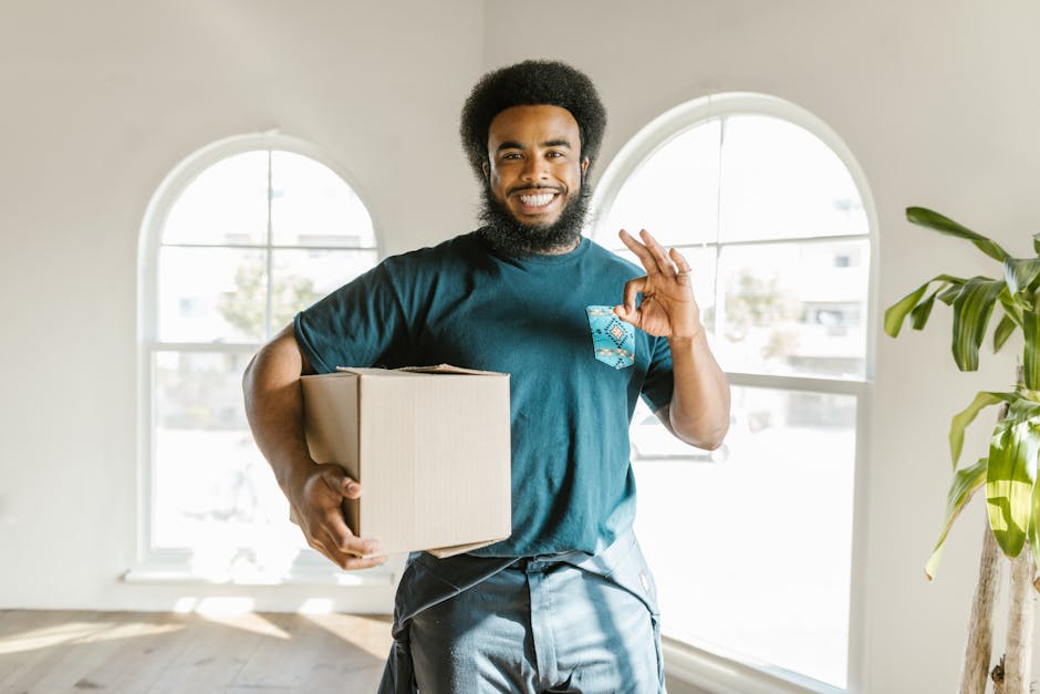 A cheerful man with an afro hairstyle and a beard is standing inside a bright, airy room with large arched windows allowing natural light to illuminate the space. He is wearing a teal t-shirt and holding a medium-sized cardboard box in his left arm, while making an 'OK' gesture with his right hand, indicating readiness for a home relocation or successful packing process. The room features light-colored walls and a green potted plant is visible to his right, adding a touch of interior decor. The scene captures a moment of packing and moving preparations, which is relevant to house removals and furniture transport services offered by Removals Haringey, as seen on the webpage about insider tips for events and moves at Alexandra Palace. The environment suggests the early stages of a household move, with the individual preparing for transportation or loading of belongings as part of a professional relocation process.