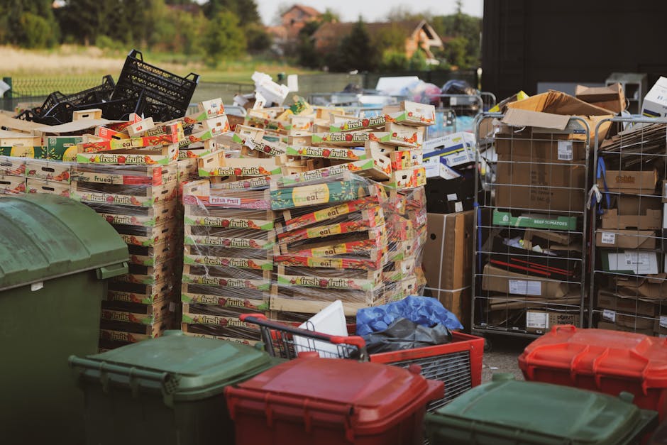 A large green outdoor waste skip filled with mixed household and packaging waste, including crumpled cardboard boxes, plastic wrap, and paper, with some cardboard pieces overflowing onto the pavement.