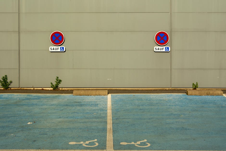 A photograph of a parking area adjacent to a beige building wall, displaying two electric vehicle charging station signs with no parking symbols, each accompanied by a blue and white wheelchair accessibility indicator and a label reading 'SAUF'. The signs are mounted on the wall and positioned evenly apart. In the foreground, there are two designated parking spaces marked with white lines and wheelchair symbols, both showing signs of wear and minor debris. The parking spaces are bordered by concrete wheel stops, and small green plants are growing along the base of the wall, close to the wheel stops. The overall environment suggests an organized parking zone possibly regulated in alignment with local traffic rules affecting parking and removals, as discussed on the related page about Haringey Council parking rules. The scene is well-lit with natural daylight, emphasizing the neutral colors of the building and the parking area.
