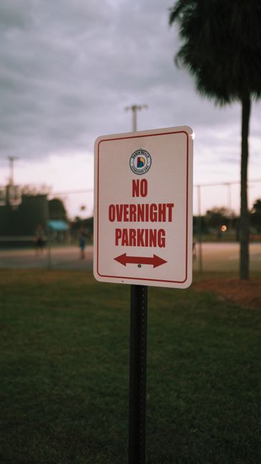 A close-up of a rectangular traffic sign with a white background and red border, displaying the message 'NO OVERNIGHT PARKING' in bold red letters, with a left and right arrow beneath the text indicating parking restrictions in both directions. The sign is mounted on a black metal post situated on a grassy area near a paved pathway. Behind the sign, there are tall palm trees with dark foliage, and a cloudy sky with a faint glow from the setting or rising sun. In the background, there is a sports field with a chain-link fence, and a few indistinct figures walking or standing near the field. The scene appears to be in an outdoor public space, and the sign's placement indicates parking regulations relevant to the vicinity, which could impact home relocation or furniture transport operations. This image exemplifies the importance of understanding local parking rules, such as those enforced by Haringey Council, during removals and moving services, and aligns with the content on removalsharingey.org.uk about how parking regulations affect booking procedures.