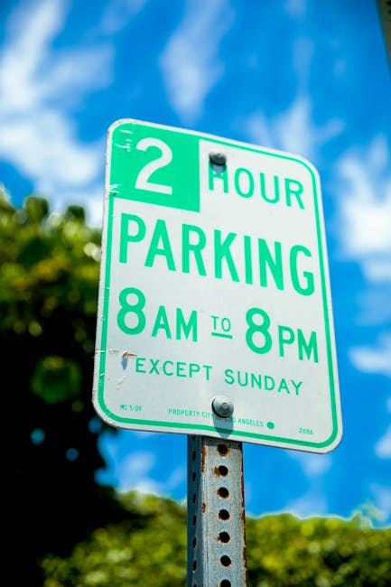 A photograph of a parking area adjacent to a beige building wall, displaying two electric vehicle charging station signs with no parking symbols, each accompanied by a blue and white wheelchair accessibility indicator and a label reading 'SAUF'. The signs are mounted on the wall and positioned evenly apart. In the foreground, there are two designated parking spaces marked with white lines and wheelchair symbols, both showing signs of wear and minor debris. The parking spaces are bordered by concrete wheel stops, and small green plants are growing along the base of the wall, close to the wheel stops. The overall environment suggests an organized parking zone possibly regulated in alignment with local traffic rules affecting parking and removals, as discussed on the related page about Haringey Council parking rules. The scene is well-lit with natural daylight, emphasizing the neutral colors of the building and the parking area.