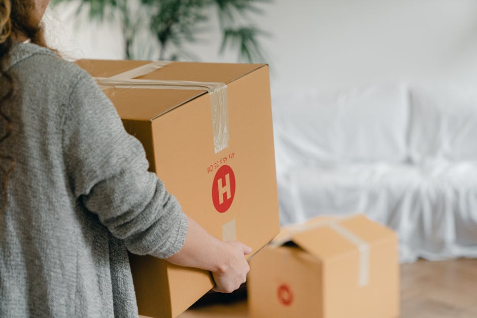A person in a grey sweater is seen holding a large cardboard moving box, with the left hand grasping the side and the right hand securing packing tape around the top. The box displays a red circular symbol with a white 'H' and is labeled with dimensions in red text. In the background, there are additional smaller cardboard boxes, some partially open, placed on the floor inside a room. The room has a white sofa covered with a white slipcover, and the lighting appears natural, coming from an unseen window, illuminating the scene. The setting suggests a home relocation process with furniture and packing materials prepared for transport, indicative of the packing and loading stages involved in house removals, which is relevant for services like those offered by Removals Haringey in the context of the N8 postcode removals cost guide on removalsharingey.org.uk.