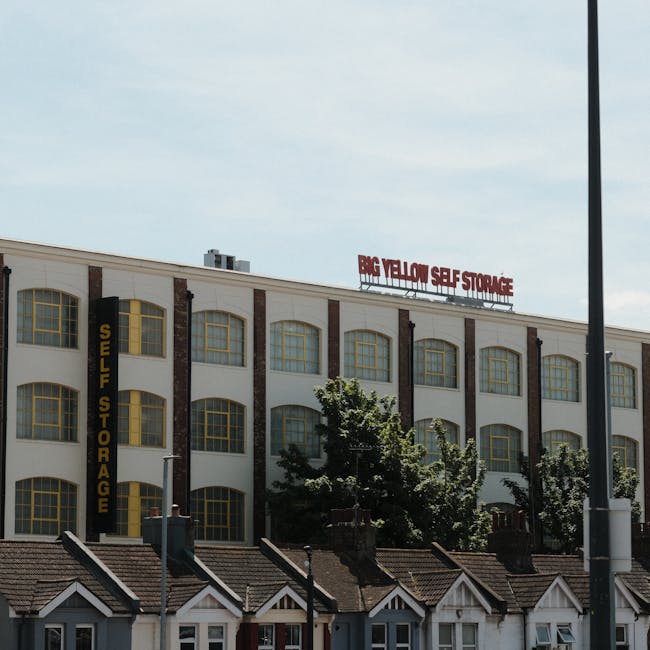 Exterior view of a large multi-storey building designated as 'Big Yellow Self Storage' with a prominent red sign on the roof indicating the storage facility's name. The building features a white facade with vertical brick accents and numerous large, arched yellow-framed windows. Mounted vertically along one side of the structure is a black sign with yellow lettering reading 'Self Storage.' In the foreground, there are small residential houses with pitched roofs and chimneys, along with a tall street lamp and some greenery, including trees partially obscuring the lower part of the storage building. The scene appears to be taken during daylight hours with partly cloudy skies, illustrating a typical commercial and residential area, relevant to home relocation, packing, and furniture transport services provided by companies like Removals Haringey.