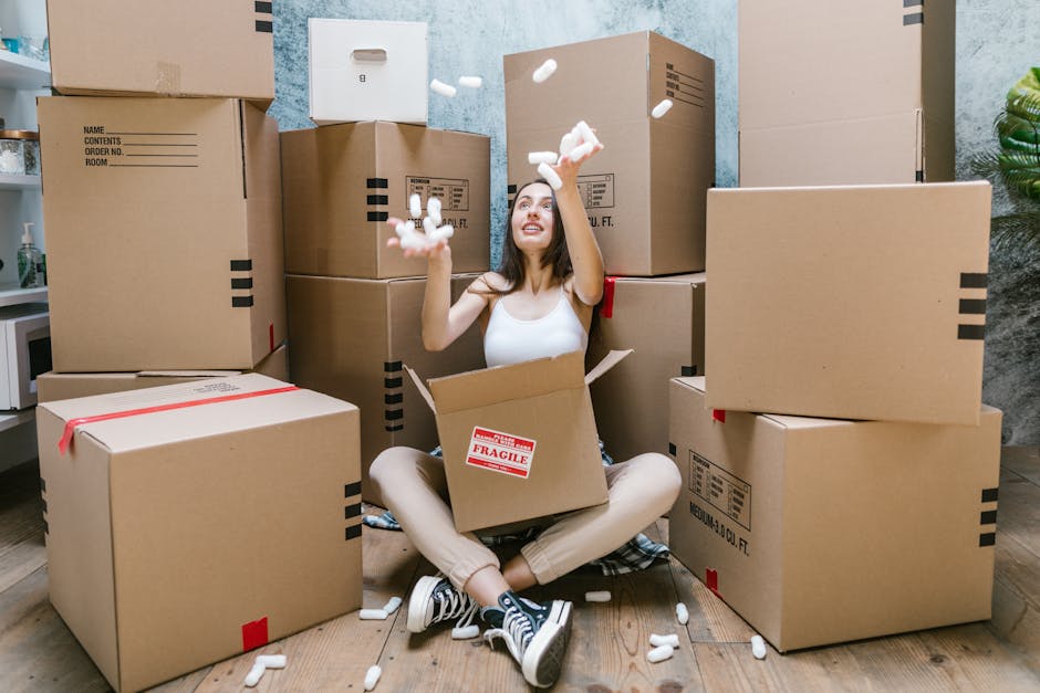 A professional removalist from Removals Haringey stands inside an empty, well-lit room with white walls and wooden flooring, holding a cardboard moving box in his right hand. The box has red and black