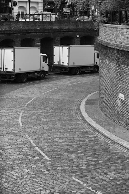 A narrow cobblestone street with a curved layout, featuring two white box trucks parked parallel to the brick wall on the right side of the image. The trucks are positioned in a way that suggests they are involved in a home relocation or furniture transport process, with the vehicles' rear doors closed. The street is bordered by a low brick wall and a larger brick retaining wall, with an arched structure or bridge visible above, possibly indicating an underground passage or tunnel. Overhead, there is a fence with trees and residential buildings in the background. The scene is in black and white, highlighting the textured cobblestones, the smooth surfaces of the trucks, and the urban environment’s structural details, as part of the logistics involved in removals and house moves. This setting is associated with ongoing loading or unloading activities typical of professional removals services such as those provided by Removals Haringey, especially considering access and lift restrictions near residential areas in Tottenham Hale.