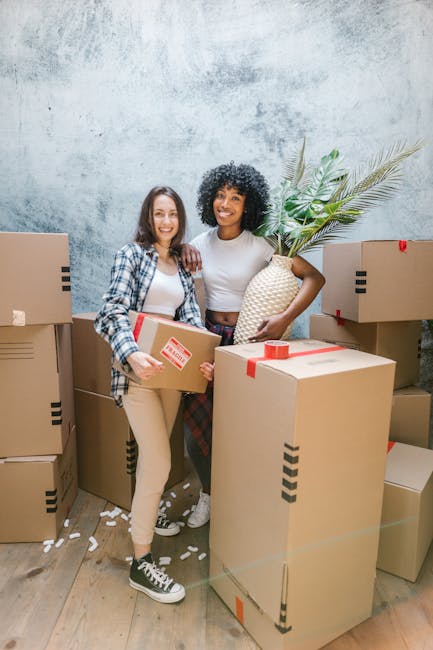 Two women standing indoors among a collection of large cardboard moving boxes, some sealed with red tape, with one woman holding an additional smaller box and the other holding a decorative woven vase with green palm leaves. The boxes are stacked around them, and there are packing materials scattered on the wooden floor. The background features a textured, light-colored wall. This scene depicts the home relocation process, including packing and moving preparations, with professional removals services such as those offered by Removals Haringey. The women are smiling at the camera, capturing a moment of assistance during a furniture transport or packing and moving activity, typical of house removals in Haringey. The environment is well-lit, highlighting the details of the packed belongings and moving supplies involved in the process.