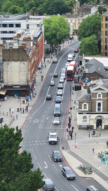 An aerial view of a busy urban street in Wood Green, with a row of parked cars along the curb and several vehicles, including a red bus, moving along the road. Pedestrians are walking on the sidewalks, some crossing at crosswalks, and a group of people is gathered near a shop frontage on the left side of the street. Mature green trees line both sides of the boulevard, providing shade in the area. Residential and commercial buildings of varying heights are visible, including a brick-style structure on the left and a smaller white building on the right. The scene captures typical city traffic and foot traffic, with a mix of moving vehicles and stationary parked cars, complemented by urban landscaping. This image reflects a typical environment where professional house removals and furniture transport might take place in Haringey, supported by local services such as Removals Haringey focusing on packing, loading, and home relocation activities.