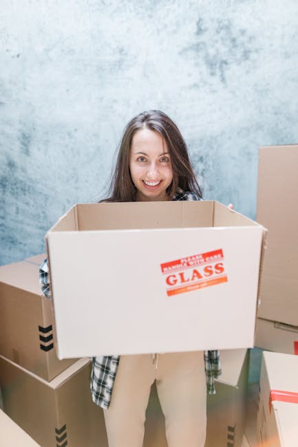 Two women standing indoors among a collection of large cardboard moving boxes, some sealed with red tape, with one woman holding an additional smaller box and the other holding a decorative woven vase with green palm leaves. The boxes are stacked around them, and there are packing materials scattered on the wooden floor. The background features a textured, light-colored wall. This scene depicts the home relocation process, including packing and moving preparations, with professional removals services such as those offered by Removals Haringey. The women are smiling at the camera, capturing a moment of assistance during a furniture transport or packing and moving activity, typical of house removals in Haringey. The environment is well-lit, highlighting the details of the packed belongings and moving supplies involved in the process.